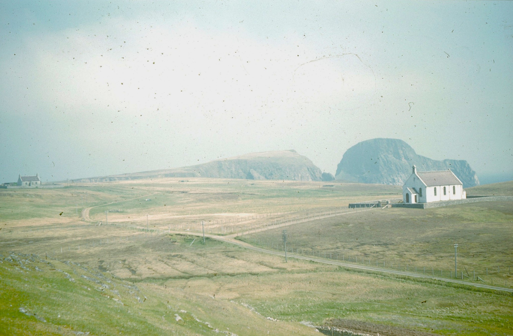 Caption: Fair Isle Church of Scotland, right, the school (now museum), far left, and Sheep Rock looming behind.